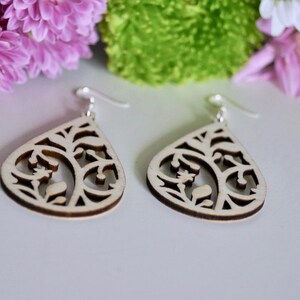 May include: A pair of wooden earrings with a delicate, intricate design. The earrings are shaped like teardrops and feature a floral pattern. The earrings are made of light-coloured wood and have silver hooks.
