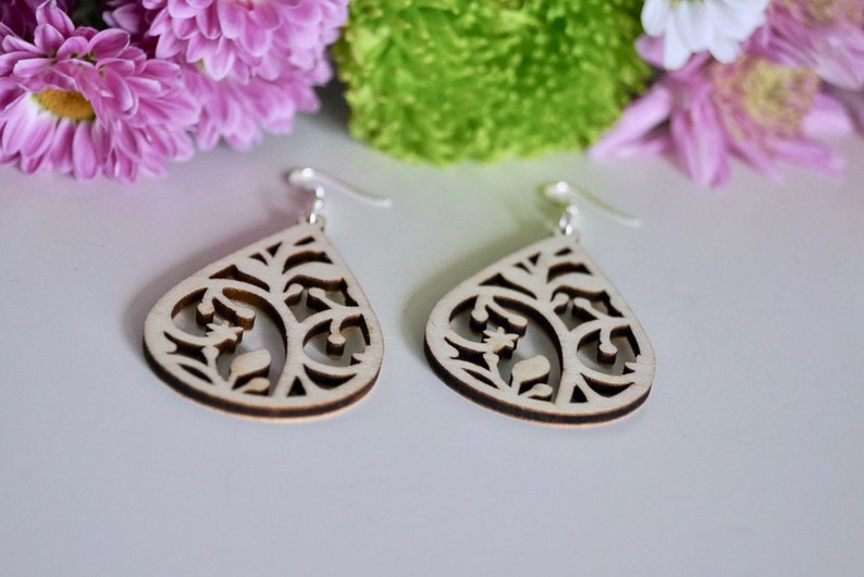 May include: A pair of wooden earrings with a delicate, intricate design. The earrings are shaped like teardrops and feature a floral pattern. The earrings are made of light-coloured wood and have silver hooks.