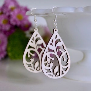 May include: A pair of white wooden earrings shaped like teardrops with a delicate floral design cut out. The earrings are hanging from silver hooks.