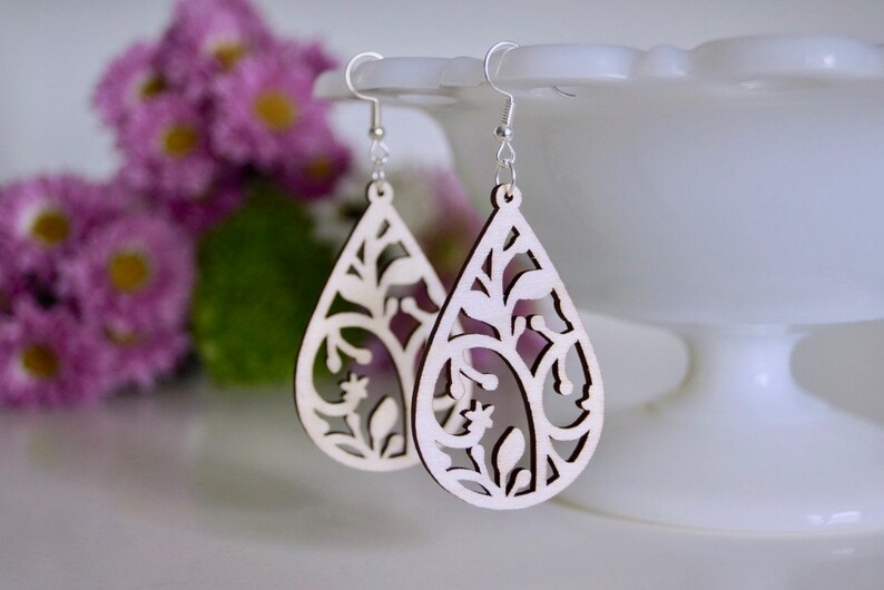 May include: A pair of white wooden earrings shaped like teardrops with a delicate floral design cut out. The earrings are hanging from silver hooks.