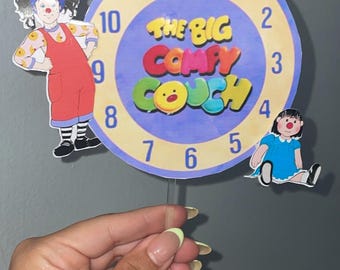 May include: A circular clock featuring the text "THE BIG COMFY COUCH" in colorful letters. The clock face has a blue background with white numbers. Cartoon characters of a clown and a girl are on either side of the clock.