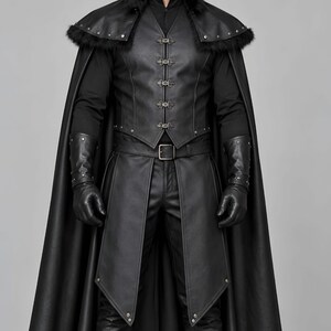 Front view of a man in a black leather wizard costume against a gray studio background. He wears a tall pointed hat, a fitted vest with silver clasps, and a long cloak with black fur trim around the collar and shoulders. Small silver rivets line the shoulder capelet and leather bracers. A buckle belt holds two long pointed waist panels over black trousers. Black gloves and tall boots complete the styling. The model faces forward with his arms relaxed, displaying the full outfit from head to toe.