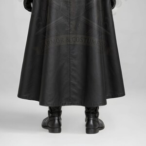 Man wearing a green leather plague doctor costume with a long lace-up coat, black buckle belt and flowing black cloak. A shoulder capelet frames the upper body, while quilted black bracers cover the forearms. The outfit is styled with black gloves, leg guards, boots, a wide-brimmed hat and a beaked mask with round eyepieces. He holds edge of the cloak and stands against a plain gray background. The vivid green coat contrasts with the black accessories, showing the costume's gothic fantasy style.