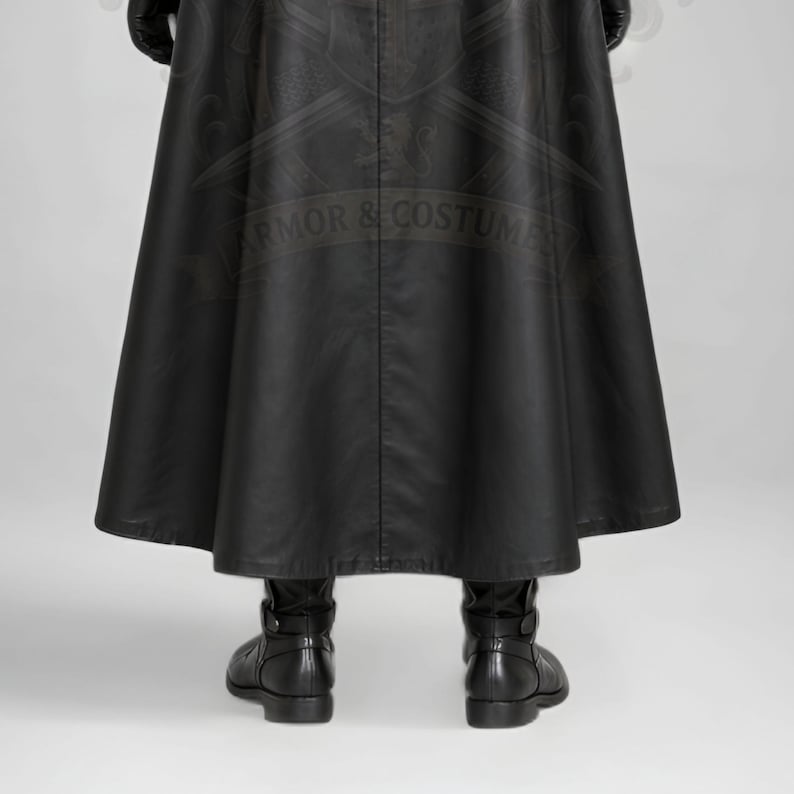 Man wearing a green leather plague doctor costume with a long lace-up coat, black buckle belt and flowing black cloak. A shoulder capelet frames the upper body, while quilted black bracers cover the forearms. The outfit is styled with black gloves, leg guards, boots, a wide-brimmed hat and a beaked mask with round eyepieces. He holds edge of the cloak and stands against a plain gray background. The vivid green coat contrasts with the black accessories, showing the costume's gothic fantasy style.