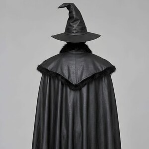 Front view of a man in a black leather wizard costume against a gray studio background. He wears a tall pointed hat, a fitted vest with silver clasps, and a long cloak with black fur trim around the collar and shoulders. Small silver rivets line the shoulder capelet and leather bracers. A buckle belt holds two long pointed waist panels over black trousers. Black gloves and tall boots complete the styling. The model faces forward with his arms relaxed, displaying the full outfit from head to toe.