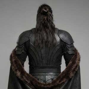 Front view of a bearded man wearing black leather Viking armor against a gray studio background. Layered angular chest panels sit above two waist buckles and long pointed leather skirt panels. A broad black cloak drapes over his shoulders, with dark brown fur edging around the collar and open front. Matching bracers cover his forearms, while strapped leg guards cover his shins over black trousers. He stands facing forward with his arms at his sides, showing the full costume and black lace boots.