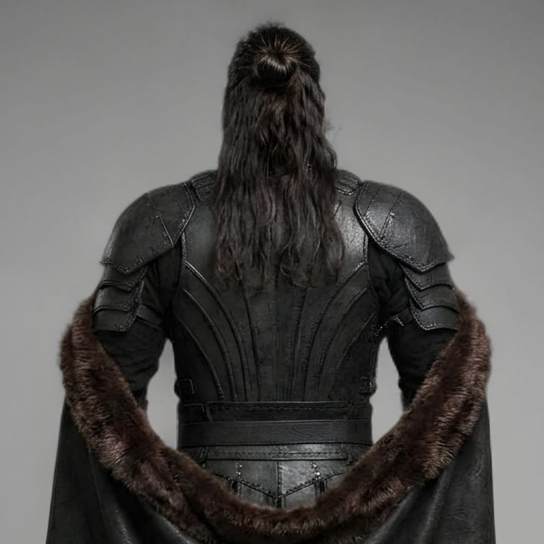 Front view of a bearded man wearing black leather Viking armor against a gray studio background. Layered angular chest panels sit above two waist buckles and long pointed leather skirt panels. A broad black cloak drapes over his shoulders, with dark brown fur edging around the collar and open front. Matching bracers cover his forearms, while strapped leg guards cover his shins over black trousers. He stands facing forward with his arms at his sides, showing the full costume and black lace boots.