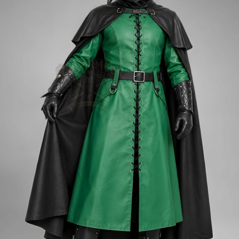 Man wearing a green leather plague doctor costume with a long lace-up coat, black buckle belt and flowing black cloak. A shoulder capelet frames the upper body, while quilted black bracers cover the forearms. The outfit is styled with black gloves, leg guards, boots, a wide-brimmed hat and a beaked mask with round eyepieces. He holds edge of the cloak and stands against a plain gray background. The vivid green coat contrasts with the black accessories, showing the costume's gothic fantasy style.