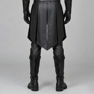 Front view of a man in a black leather wizard costume against a gray studio background. He wears a tall pointed hat, a fitted vest with silver clasps, and a long cloak with black fur trim around the collar and shoulders. Small silver rivets line the shoulder capelet and leather bracers. A buckle belt holds two long pointed waist panels over black trousers. Black gloves and tall boots complete the styling. The model faces forward with his arms relaxed, displaying the full outfit from head to toe.