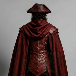 Men's leather pirate costume in dark red and black, featuring a detailed leather vest, layered shoulder armor, arm bracers, leg armor, and flowing cape. Handmade pirate cosplay outfit for Halloween, LARP, Renaissance fairs, medieval fantasy events, themed parties, festivals, conventions, stage performances, and photoshoots. Custom sizing, colors, length, and design options available. Dark fantasy pirate captain costume with wearable leather armor and handcrafted details.