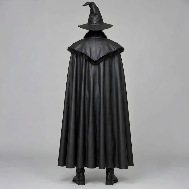 Front view of a man in a black leather wizard costume against a gray studio background. He wears a tall pointed hat, a fitted vest with silver clasps, and a long cloak with black fur trim around the collar and shoulders. Small silver rivets line the shoulder capelet and leather bracers. A buckle belt holds two long pointed waist panels over black trousers. Black gloves and tall boots complete the styling. The model faces forward with his arms relaxed, displaying the full outfit from head to toe.