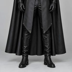 Front view of a man in a black leather wizard costume against a gray studio background. He wears a tall pointed hat, a fitted vest with silver clasps, and a long cloak with black fur trim around the collar and shoulders. Small silver rivets line the shoulder capelet and leather bracers. A buckle belt holds two long pointed waist panels over black trousers. Black gloves and tall boots complete the styling. The model faces forward with his arms relaxed, displaying the full outfit from head to toe.