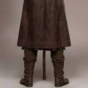 Full-length studio portrait of a rugged Viking warrior standing against a soft gray background. He wears a long brown leather coat with a wide fur-trimmed collar and fur edging down the front, layered over a matching leather tunic with toggle closures. Leather bracers, fitted pants, and tall wrapped boots complete the look. The bald, bearded man stands firmly with both hands resting on a wooden staff, creating a bold medieval Norse costume appearance.