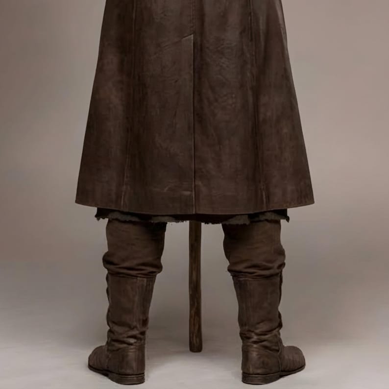 Full-length studio portrait of a rugged Viking warrior standing against a soft gray background. He wears a long brown leather coat with a wide fur-trimmed collar and fur edging down the front, layered over a matching leather tunic with toggle closures. Leather bracers, fitted pants, and tall wrapped boots complete the look. The bald, bearded man stands firmly with both hands resting on a wooden staff, creating a bold medieval Norse costume appearance.