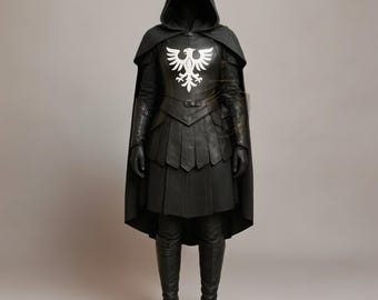 Puede incluir: Disfraz de cuero negro con capa con capucha. El disfraz presenta un emblema de águila blanca en el pecho, una falda plisada y botas altas hasta la rodilla. El atuendo incluye guantes y una capa.