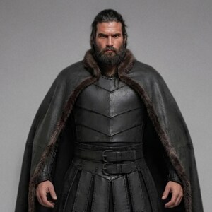 Front view of a bearded man wearing black leather Viking armor against a gray studio background. Layered angular chest panels sit above two waist buckles and long pointed leather skirt panels. A broad black cloak drapes over his shoulders, with dark brown fur edging around the collar and open front. Matching bracers cover his forearms, while strapped leg guards cover his shins over black trousers. He stands facing forward with his arms at his sides, showing the full costume and black lace boots.