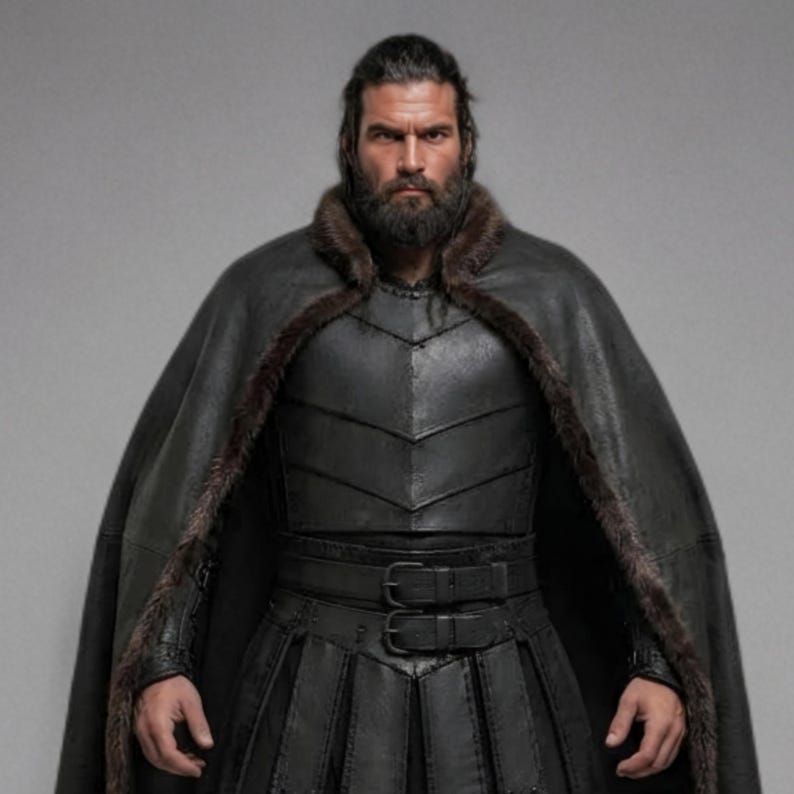 Front view of a bearded man wearing black leather Viking armor against a gray studio background. Layered angular chest panels sit above two waist buckles and long pointed leather skirt panels. A broad black cloak drapes over his shoulders, with dark brown fur edging around the collar and open front. Matching bracers cover his forearms, while strapped leg guards cover his shins over black trousers. He stands facing forward with his arms at his sides, showing the full costume and black lace boots.