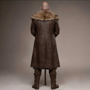 Full-length studio portrait of a rugged Viking warrior standing against a soft gray background. He wears a long brown leather coat with a wide fur-trimmed collar and fur edging down the front, layered over a matching leather tunic with toggle closures. Leather bracers, fitted pants, and tall wrapped boots complete the look. The bald, bearded man stands firmly with both hands resting on a wooden staff, creating a bold medieval Norse costume appearance.