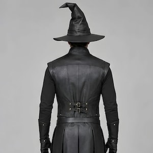 Front view of a man in a black leather wizard costume against a gray studio background. He wears a tall pointed hat, a fitted vest with silver clasps, and a long cloak with black fur trim around the collar and shoulders. Small silver rivets line the shoulder capelet and leather bracers. A buckle belt holds two long pointed waist panels over black trousers. Black gloves and tall boots complete the styling. The model faces forward with his arms relaxed, displaying the full outfit from head to toe.