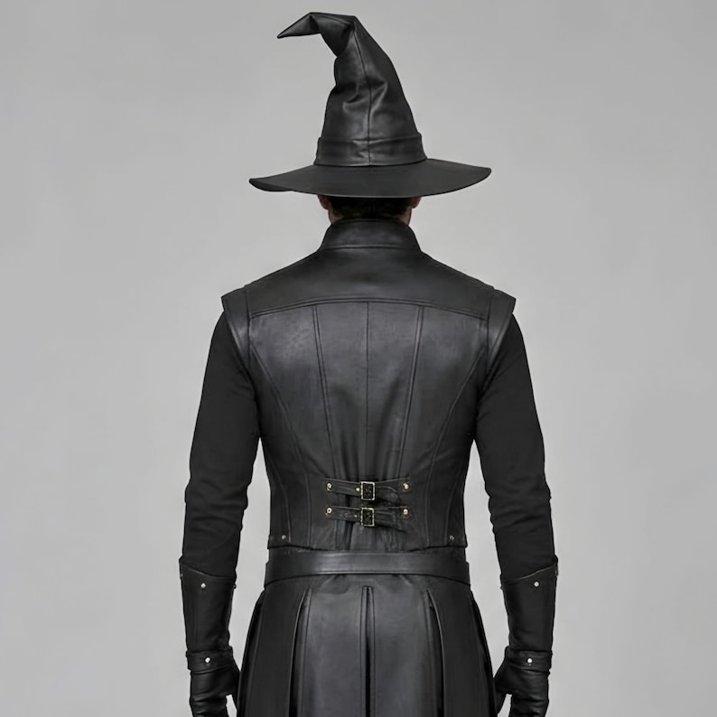 Front view of a man in a black leather wizard costume against a gray studio background. He wears a tall pointed hat, a fitted vest with silver clasps, and a long cloak with black fur trim around the collar and shoulders. Small silver rivets line the shoulder capelet and leather bracers. A buckle belt holds two long pointed waist panels over black trousers. Black gloves and tall boots complete the styling. The model faces forward with his arms relaxed, displaying the full outfit from head to toe.