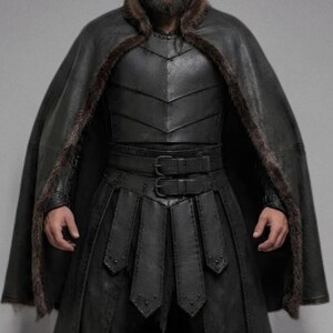 Front view of a bearded man wearing black leather Viking armor against a gray studio background. Layered angular chest panels sit above two waist buckles and long pointed leather skirt panels. A broad black cloak drapes over his shoulders, with dark brown fur edging around the collar and open front. Matching bracers cover his forearms, while strapped leg guards cover his shins over black trousers. He stands facing forward with his arms at his sides, showing the full costume and black lace boots.