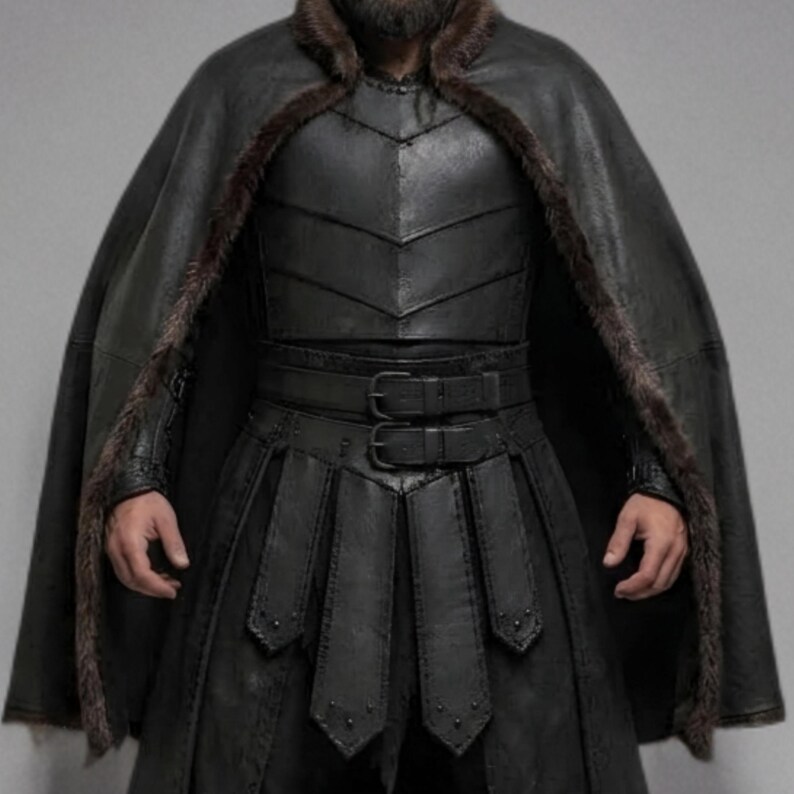 Front view of a bearded man wearing black leather Viking armor against a gray studio background. Layered angular chest panels sit above two waist buckles and long pointed leather skirt panels. A broad black cloak drapes over his shoulders, with dark brown fur edging around the collar and open front. Matching bracers cover his forearms, while strapped leg guards cover his shins over black trousers. He stands facing forward with his arms at his sides, showing the full costume and black lace boots.