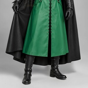 Man wearing a green leather plague doctor costume with a long lace-up coat, black buckle belt and flowing black cloak. A shoulder capelet frames the upper body, while quilted black bracers cover the forearms. The outfit is styled with black gloves, leg guards, boots, a wide-brimmed hat and a beaked mask with round eyepieces. He holds edge of the cloak and stands against a plain gray background. The vivid green coat contrasts with the black accessories, showing the costume's gothic fantasy style.