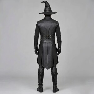 Front view of a man in a black leather wizard costume against a gray studio background. He wears a tall pointed hat, a fitted vest with silver clasps, and a long cloak with black fur trim around the collar and shoulders. Small silver rivets line the shoulder capelet and leather bracers. A buckle belt holds two long pointed waist panels over black trousers. Black gloves and tall boots complete the styling. The model faces forward with his arms relaxed, displaying the full outfit from head to toe.