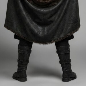 Front view of a bearded man wearing black leather Viking armor against a gray studio background. Layered angular chest panels sit above two waist buckles and long pointed leather skirt panels. A broad black cloak drapes over his shoulders, with dark brown fur edging around the collar and open front. Matching bracers cover his forearms, while strapped leg guards cover his shins over black trousers. He stands facing forward with his arms at his sides, showing the full costume and black lace boots.