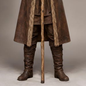 Full-length studio portrait of a rugged Viking warrior standing against a soft gray background. He wears a long brown leather coat with a wide fur-trimmed collar and fur edging down the front, layered over a matching leather tunic with toggle closures. Leather bracers, fitted pants, and tall wrapped boots complete the look. The bald, bearded man stands firmly with both hands resting on a wooden staff, creating a bold medieval Norse costume appearance.