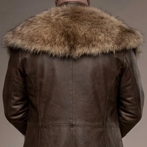 Full-length studio portrait of a rugged Viking warrior standing against a soft gray background. He wears a long brown leather coat with a wide fur-trimmed collar and fur edging down the front, layered over a matching leather tunic with toggle closures. Leather bracers, fitted pants, and tall wrapped boots complete the look. The bald, bearded man stands firmly with both hands resting on a wooden staff, creating a bold medieval Norse costume appearance.