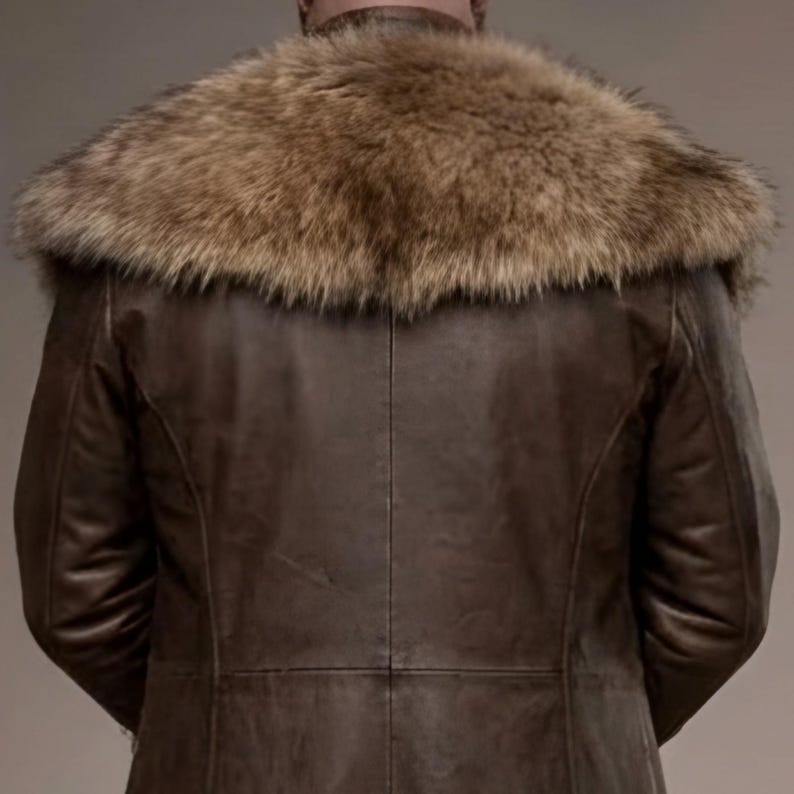 Full-length studio portrait of a rugged Viking warrior standing against a soft gray background. He wears a long brown leather coat with a wide fur-trimmed collar and fur edging down the front, layered over a matching leather tunic with toggle closures. Leather bracers, fitted pants, and tall wrapped boots complete the look. The bald, bearded man stands firmly with both hands resting on a wooden staff, creating a bold medieval Norse costume appearance.
