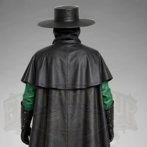 Man wearing a green leather plague doctor costume with a long lace-up coat, black buckle belt and flowing black cloak. A shoulder capelet frames the upper body, while quilted black bracers cover the forearms. The outfit is styled with black gloves, leg guards, boots, a wide-brimmed hat and a beaked mask with round eyepieces. He holds edge of the cloak and stands against a plain gray background. The vivid green coat contrasts with the black accessories, showing the costume's gothic fantasy style.