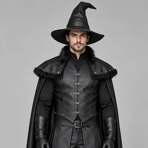 Front view of a man in a black leather wizard costume against a gray studio background. He wears a tall pointed hat, a fitted vest with silver clasps, and a long cloak with black fur trim around the collar and shoulders. Small silver rivets line the shoulder capelet and leather bracers. A buckle belt holds two long pointed waist panels over black trousers. Black gloves and tall boots complete the styling. The model faces forward with his arms relaxed, displaying the full outfit from head to toe.