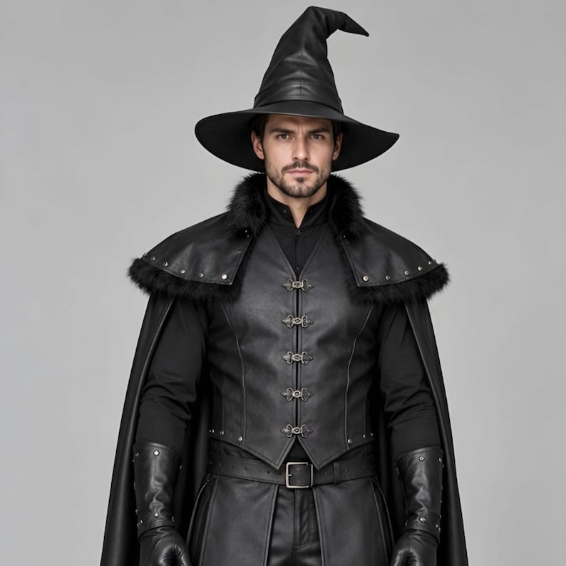 Front view of a man in a black leather wizard costume against a gray studio background. He wears a tall pointed hat, a fitted vest with silver clasps, and a long cloak with black fur trim around the collar and shoulders. Small silver rivets line the shoulder capelet and leather bracers. A buckle belt holds two long pointed waist panels over black trousers. Black gloves and tall boots complete the styling. The model faces forward with his arms relaxed, displaying the full outfit from head to toe.