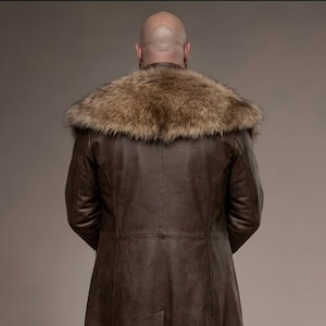 Full-length studio portrait of a rugged Viking warrior standing against a soft gray background. He wears a long brown leather coat with a wide fur-trimmed collar and fur edging down the front, layered over a matching leather tunic with toggle closures. Leather bracers, fitted pants, and tall wrapped boots complete the look. The bald, bearded man stands firmly with both hands resting on a wooden staff, creating a bold medieval Norse costume appearance.