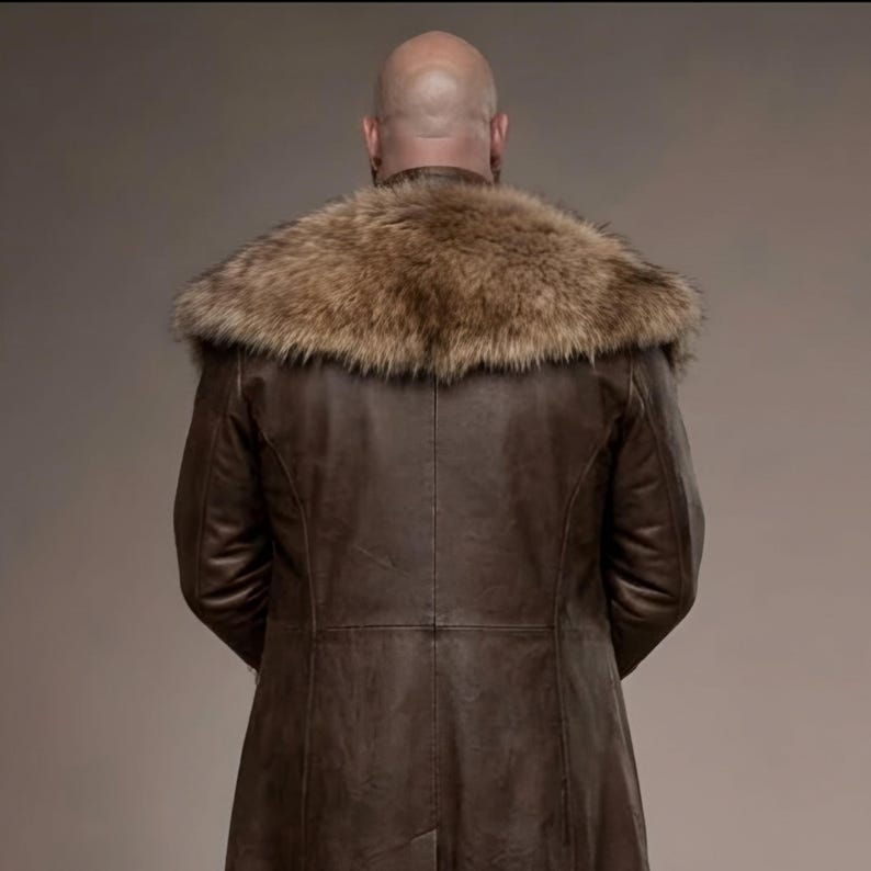 Full-length studio portrait of a rugged Viking warrior standing against a soft gray background. He wears a long brown leather coat with a wide fur-trimmed collar and fur edging down the front, layered over a matching leather tunic with toggle closures. Leather bracers, fitted pants, and tall wrapped boots complete the look. The bald, bearded man stands firmly with both hands resting on a wooden staff, creating a bold medieval Norse costume appearance.