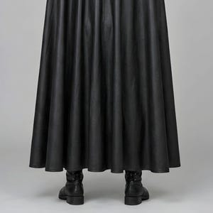 Front view of a man in a black leather wizard costume against a gray studio background. He wears a tall pointed hat, a fitted vest with silver clasps, and a long cloak with black fur trim around the collar and shoulders. Small silver rivets line the shoulder capelet and leather bracers. A buckle belt holds two long pointed waist panels over black trousers. Black gloves and tall boots complete the styling. The model faces forward with his arms relaxed, displaying the full outfit from head to toe.