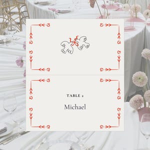 May include: Ivory place card with red heart and arrow border, featuring a line drawing of two doves tied with a ribbon. The card reads "TABLE 2" and "Michael" in a dark blue font. The card is set on a white tablecloth.