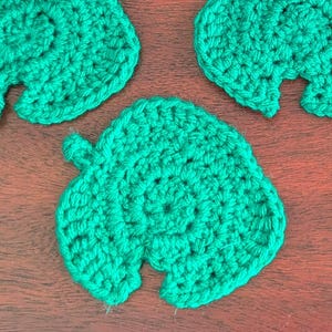 May include: Five handmade, green crochet leaf-shaped coasters. The coasters are arranged on a dark wooden surface. Each coaster has a detailed, textured design, perfect for adding a touch of nature to your home decor.