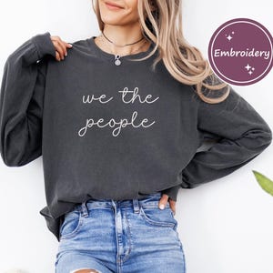 May include: A dark gray long-sleeve shirt with the words "we the people" in white script. The shirt is paired with blue jeans. A circular "Embroidery" graphic is in the upper right corner.