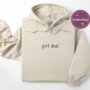 May include: A beige hoodie with the text "girl dad" in black. The hoodie has a drawstring hood and long sleeves. A circular "Embroidery" sticker is in the upper right corner. The hoodie is made of a soft, comfortable material.