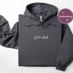 Puede incluir: Sudadera con capucha gris oscuro con el texto "girl dad" en blanco. La sudadera tiene una capucha con cord&oacute;n y un bolsillo de canguro. La palabra "Embroidery" est&aacute; en un c&iacute;rculo morado en la esquina superior derecha.
