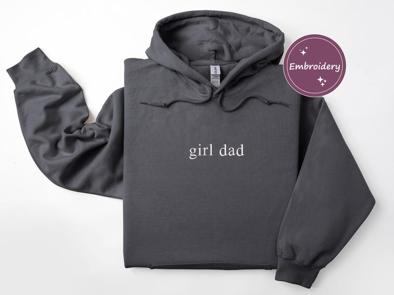 Puede incluir: Sudadera con capucha gris oscuro con el texto "girl dad" en blanco. La sudadera tiene una capucha con cord&oacute;n y un bolsillo de canguro. La palabra "Embroidery" est&aacute; en un c&iacute;rculo morado en la esquina superior derecha.
