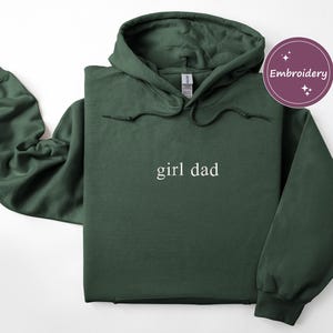 Puede incluir: Sudadera con capucha verde oscuro con el texto "girl dad" en blanco. La sudadera tiene una capucha con cord&oacute;n y un bolsillo de canguro. La palabra "Embroidery" est&aacute; en un c&iacute;rculo morado en la esquina superior derecha.