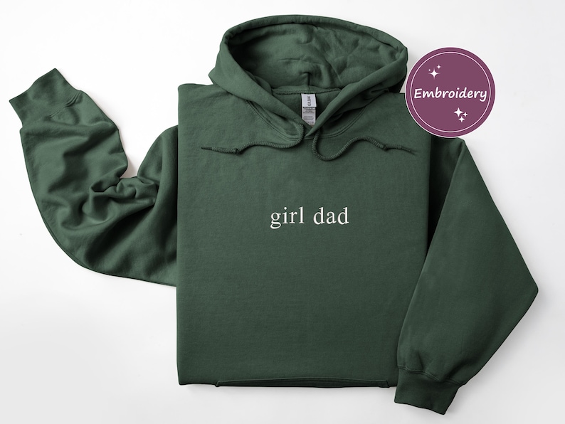Puede incluir: Sudadera con capucha verde oscuro con el texto "girl dad" en blanco. La sudadera tiene una capucha con cord&oacute;n y un bolsillo de canguro. La palabra "Embroidery" est&aacute; en un c&iacute;rculo morado en la esquina superior derecha.