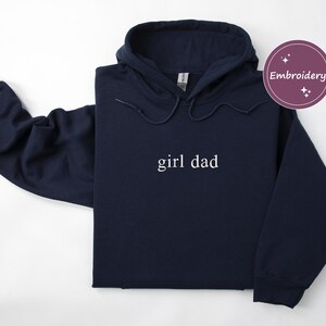 Puede incluir: Una sudadera con capucha azul marino con las palabras "girl dad" en letras blancas. La sudadera tiene una capucha con cord&oacute;n y mangas largas. Tambi&eacute;n se ve un c&iacute;rculo morado con la palabra "Bordado".