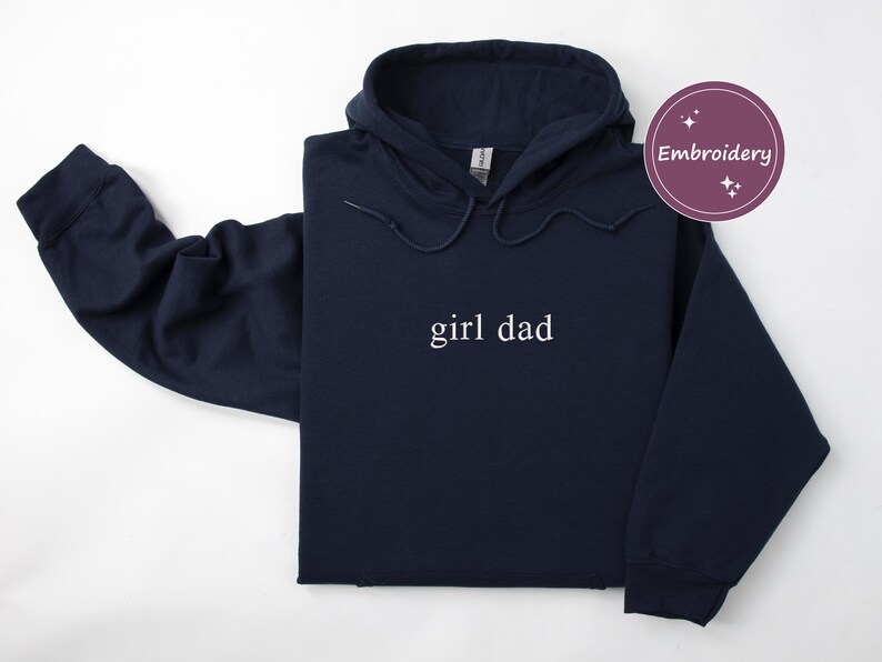 Puede incluir: Una sudadera con capucha azul marino con las palabras "girl dad" en letras blancas. La sudadera tiene una capucha con cord&oacute;n y mangas largas. Tambi&eacute;n se ve un c&iacute;rculo morado con la palabra "Bordado".