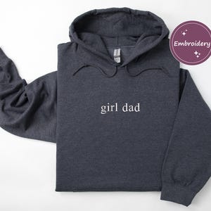 Puede incluir: Sudadera con capucha gris oscuro con el texto "girl dad" en blanco. La sudadera tiene una capucha con cord&oacute;n y mangas largas. Un c&iacute;rculo p&uacute;rpura con la palabra "Embroidery" est&aacute; en la esquina superior derecha.