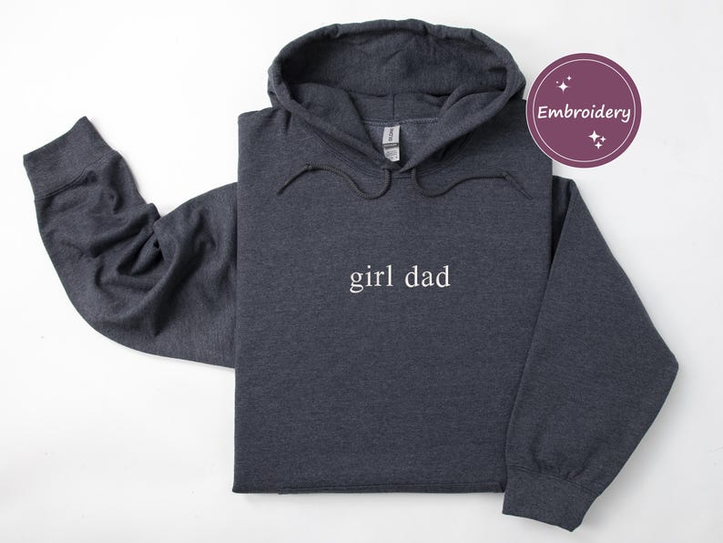Puede incluir: Sudadera con capucha gris oscuro con el texto "girl dad" en blanco. La sudadera tiene una capucha con cord&oacute;n y mangas largas. Un c&iacute;rculo p&uacute;rpura con la palabra "Embroidery" est&aacute; en la esquina superior derecha.